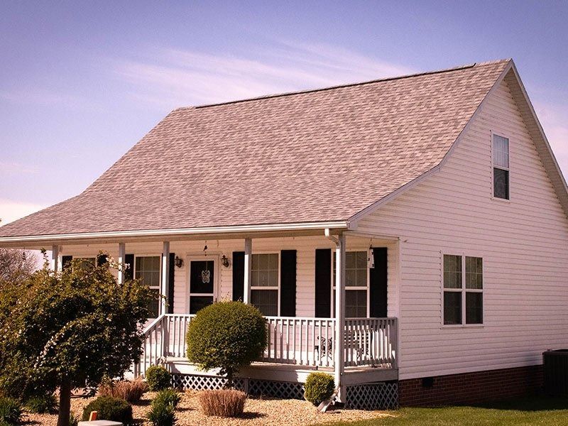 White house with a porch, gray roof, and black shutters, in a sunny setting.