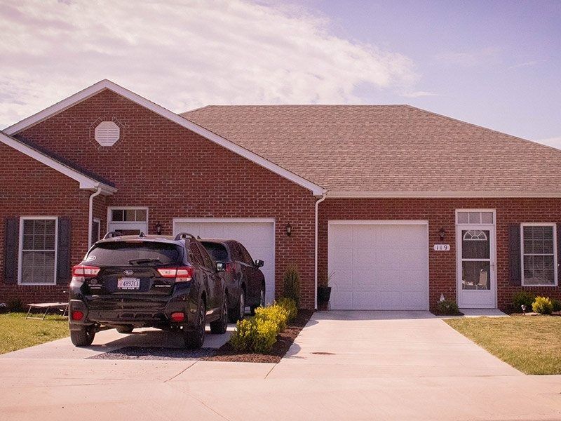 A brick house with a two-car garage, driveway, and SUV parked in front.