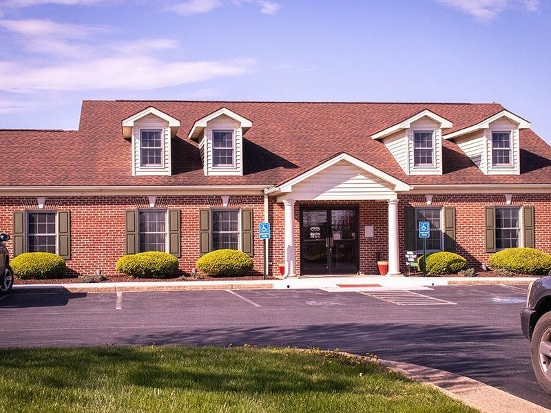 Brick building with red roof and three dormers, accessible entrance, green bushes, and a parking lot.