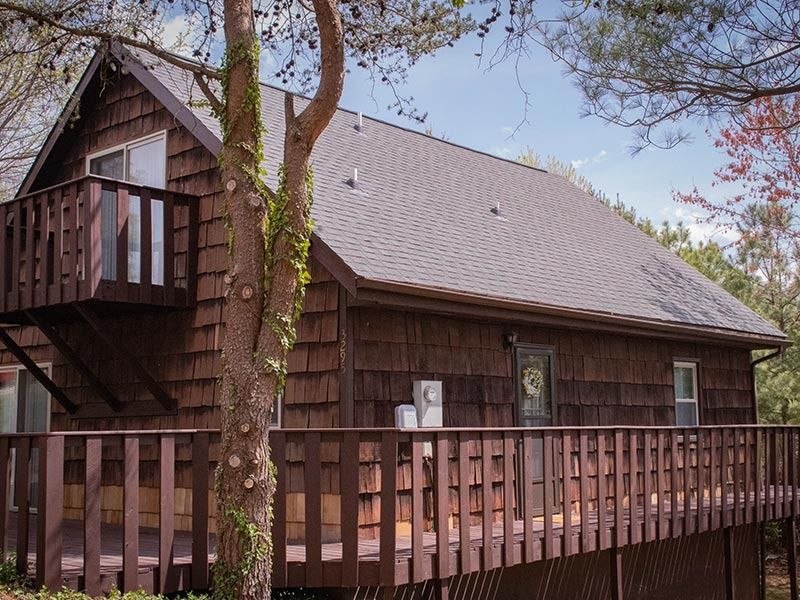 Wooden cabin with brown shingles, deck, and balcony, set among trees.