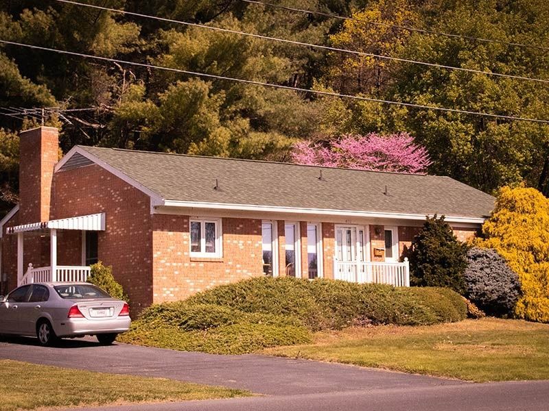 Brick ranch house with car in driveway, surrounded by trees and bushes.