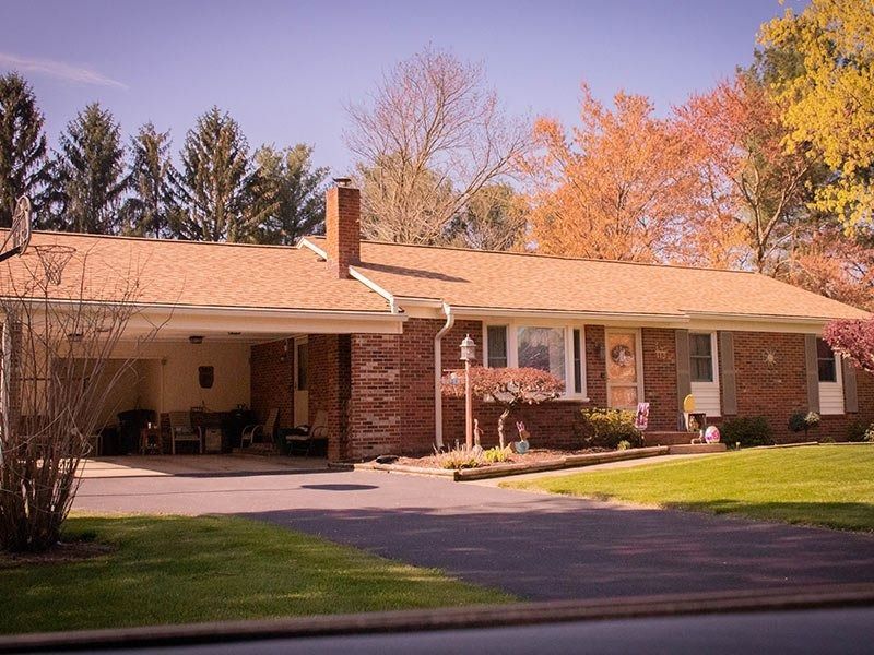 Brick ranch-style house with carport, driveway, and lawn; trees in the background; sunny day.