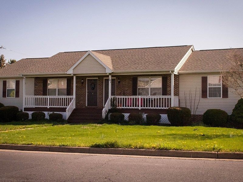 Ranch-style house with tan siding, brown roof, and porch. Front yard with green grass.