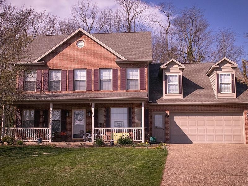 Two-story brick house with brown roof, front porch, and two dormer windows, set in a yard with trees.