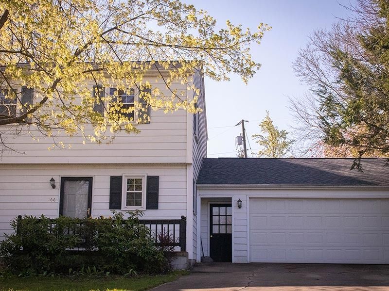 White two-story house with attached garage; black door and shutters; green bushes; tree with yellow leaves.