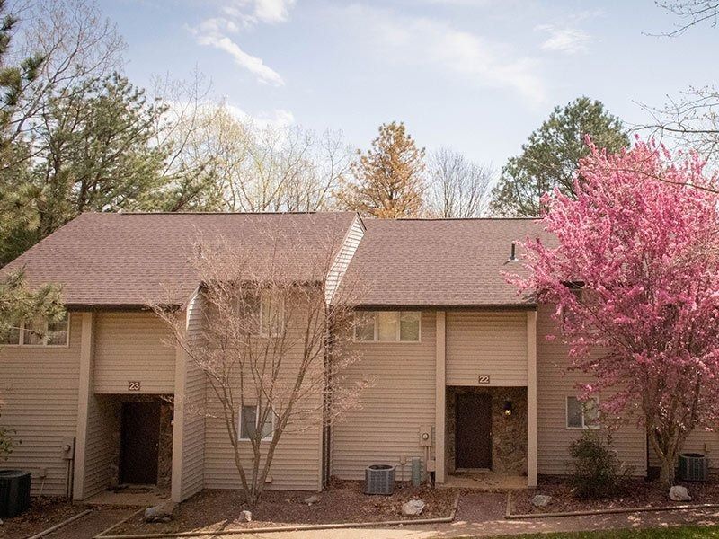 Townhouses with beige siding and brown roof under a blue sky, trees surround.