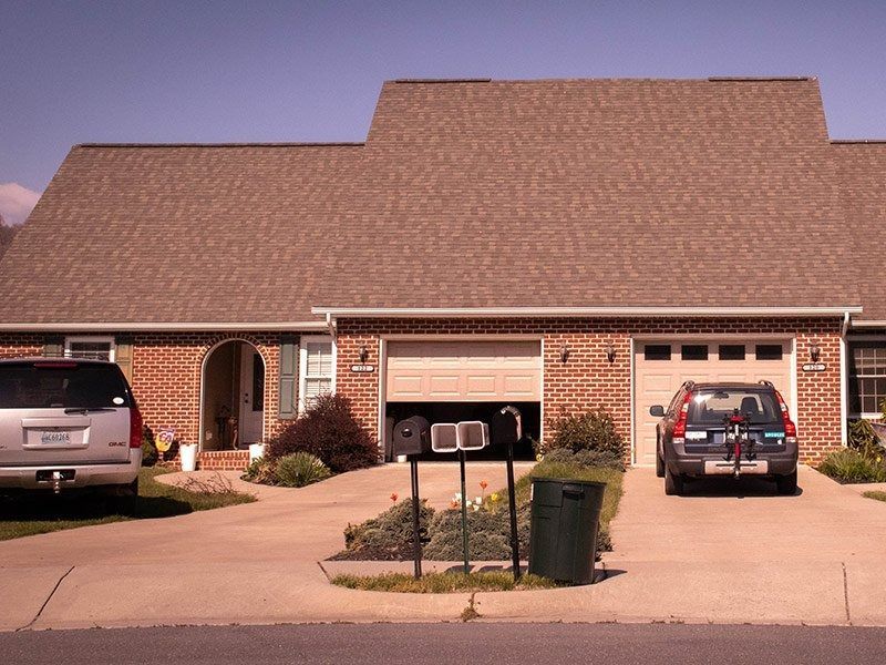 Brick duplex with two garages, driveways, and vehicles parked in them. Mailbox and trash can in front.