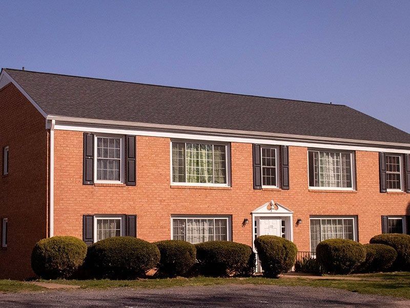 Two-story brick building with black shutters, dark roof, and manicured bushes against a clear blue sky.
