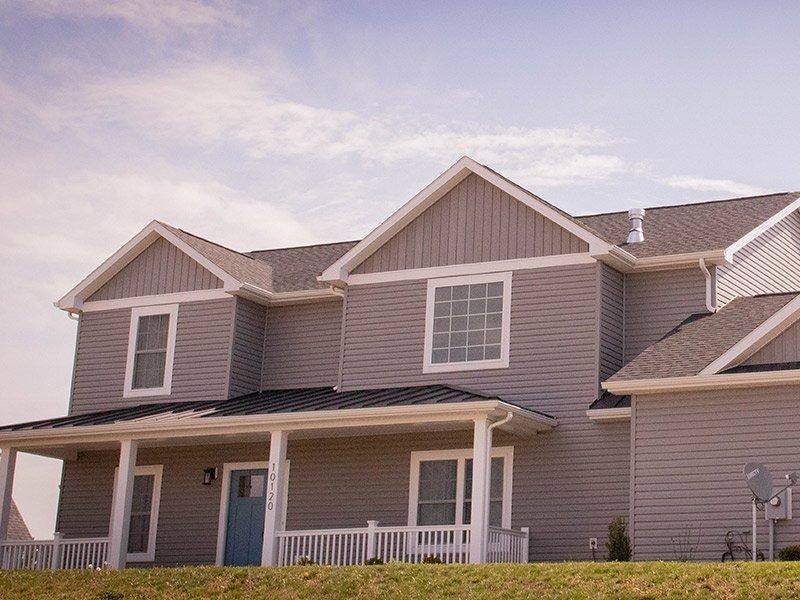 Two-story gray house with white trim, porch, and blue door, on a grassy hill.