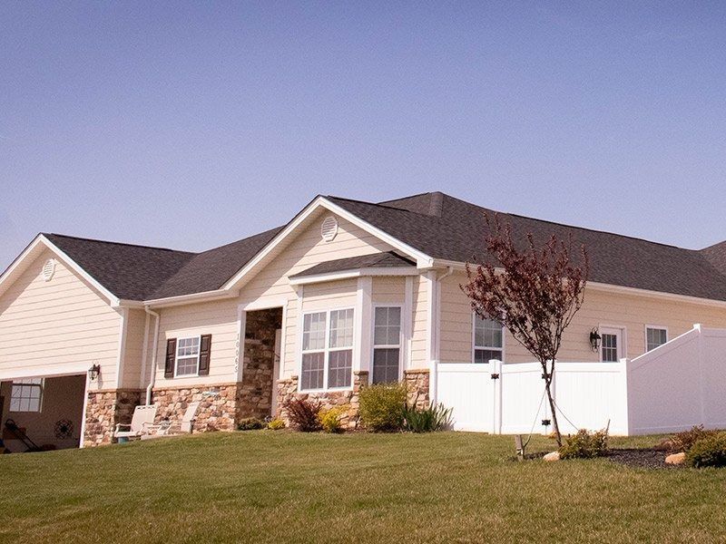 Beige house with stone accents, bay window, black shutters, and white fence on a green lawn under a blue sky.