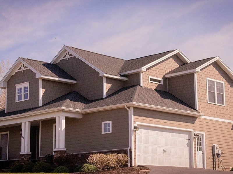 Tan two-story house with a white garage door, porch, and decorative roof accents against a blue sky.