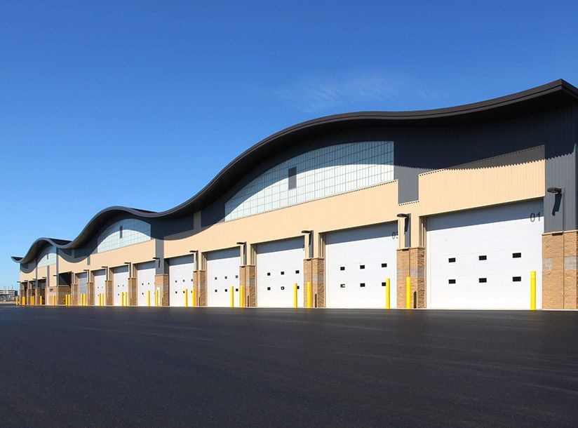 Tan industrial building with wavy roofline, multiple garage doors, and a clear blue sky.