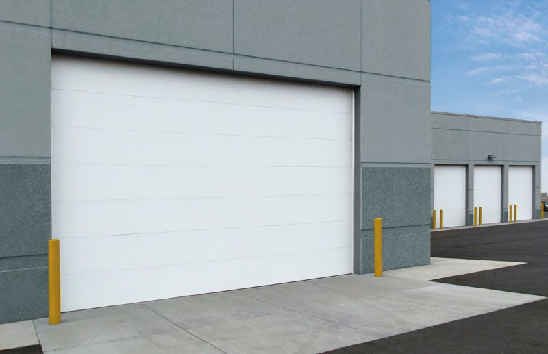 White overhead door on a gray building with yellow bollards; other similar doors in the background.