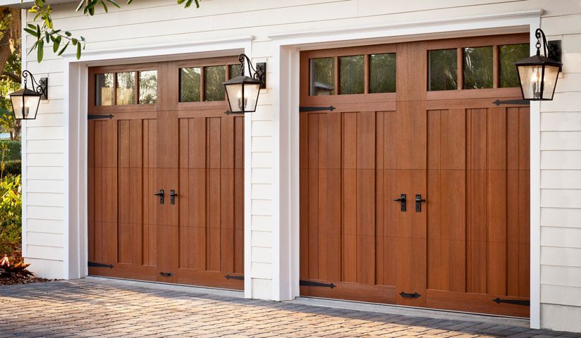 Two wooden garage doors with windows, flanked by lanterns, set against a white wall and a brick driveway.