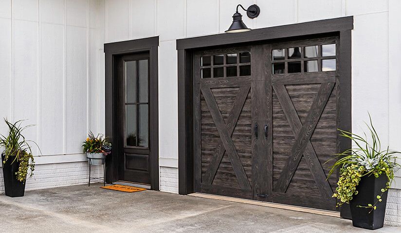 Dark wood garage doors with sidelight and potted plants against a white building exterior.