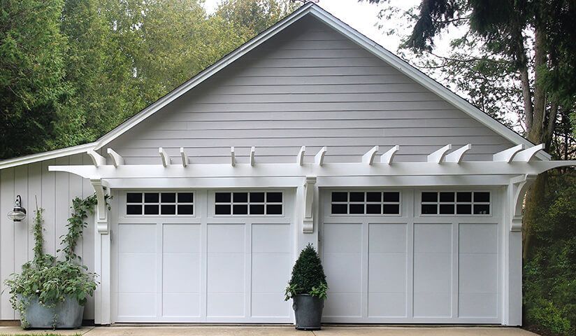 White garage with two doors, pergola above. Gray siding, greenery, and trees in background.