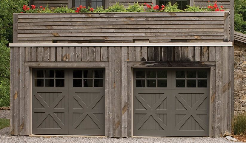 Two gray garage doors with decorative cross patterns, set in a rustic wooden structure with a plant box on top.