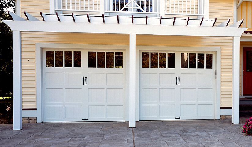 White garage doors under a white pergola, set against a yellow building with a concrete driveway.