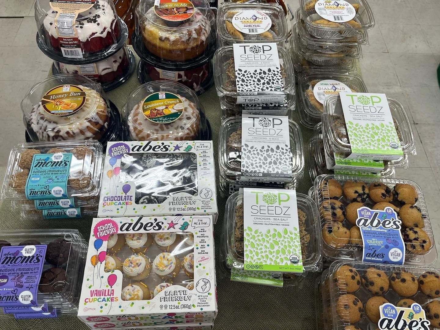 Bakery display: cakes, cupcakes, and cookies in clear plastic containers, various brands and flavors.