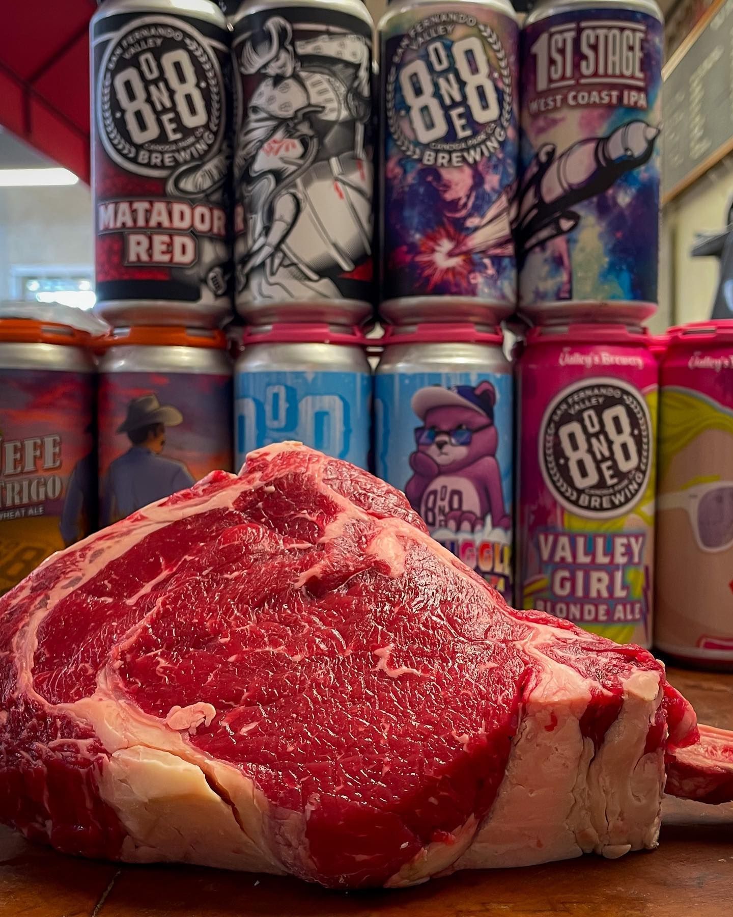 A large raw steak with beer cans stacked in the background. The cans are various colors and labeled "80one Brewery".