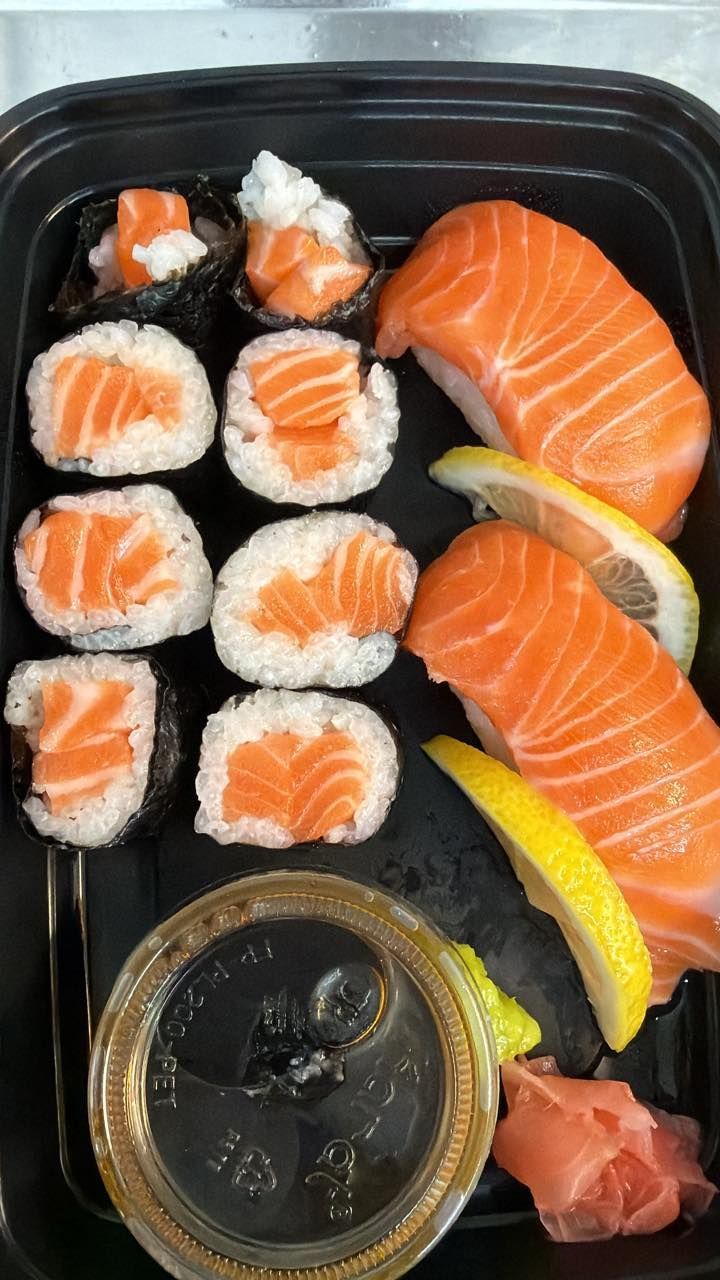 A plastic container filled with salmon sushi rolls, two pieces of salmon nigiri, soy sauce, pickled ginger, and lemon slices.