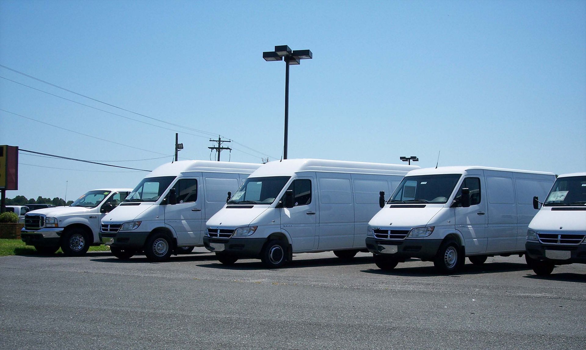 A row of white cargo vans parked on an asphalt lot under a bright, clear blue sky.
