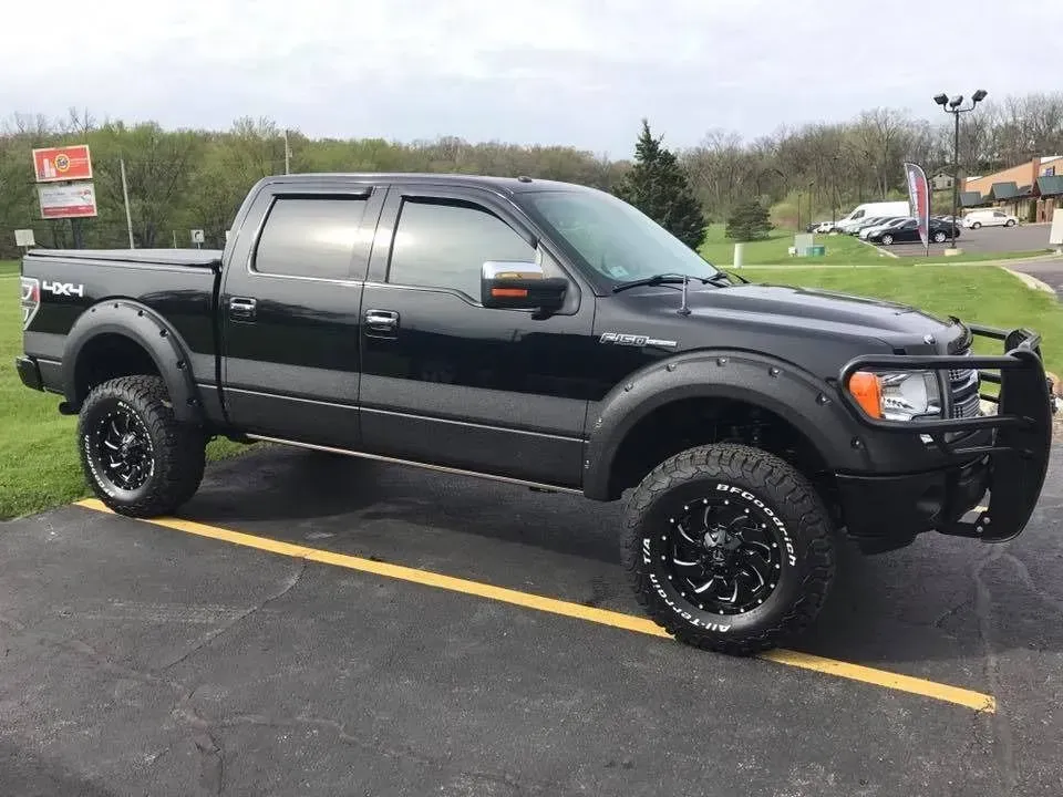 Black Ford F-150 truck with large wheels and brush guard parked on asphalt, cloudy day.