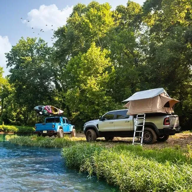 Two trucks parked by a river, one with a rooftop tent, another with kayaks, near trees on a sunny day.