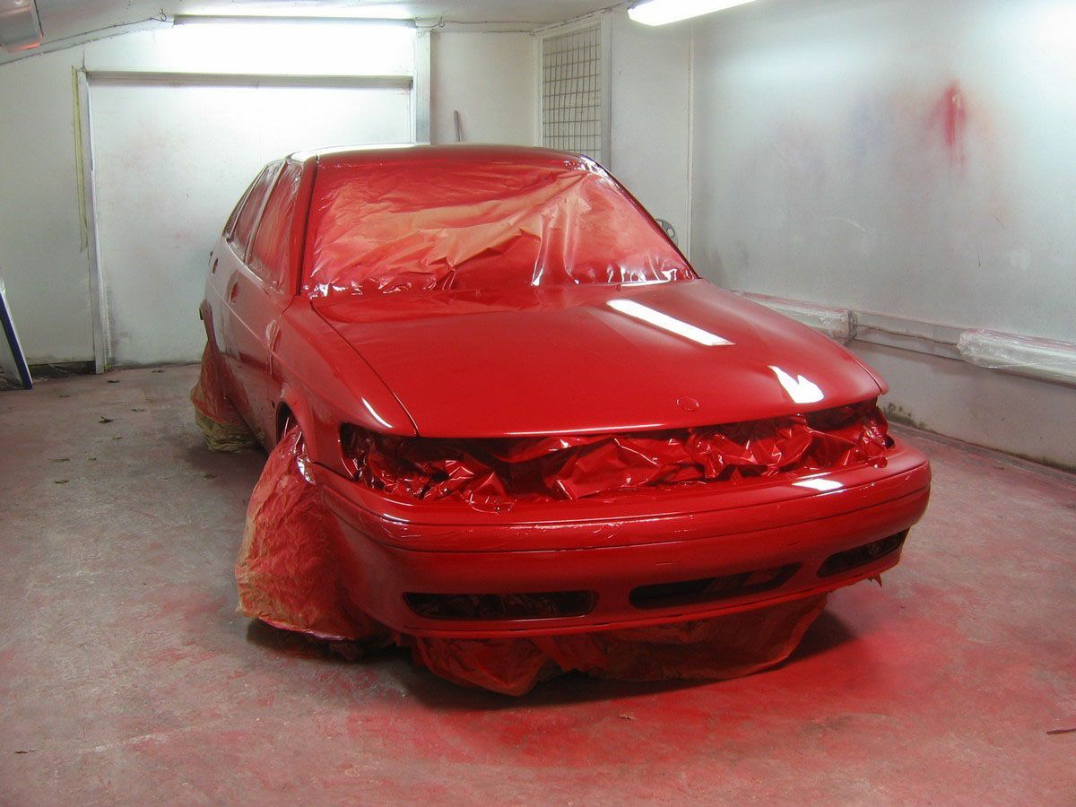 Red car covered in plastic sheeting inside a paint booth or garage