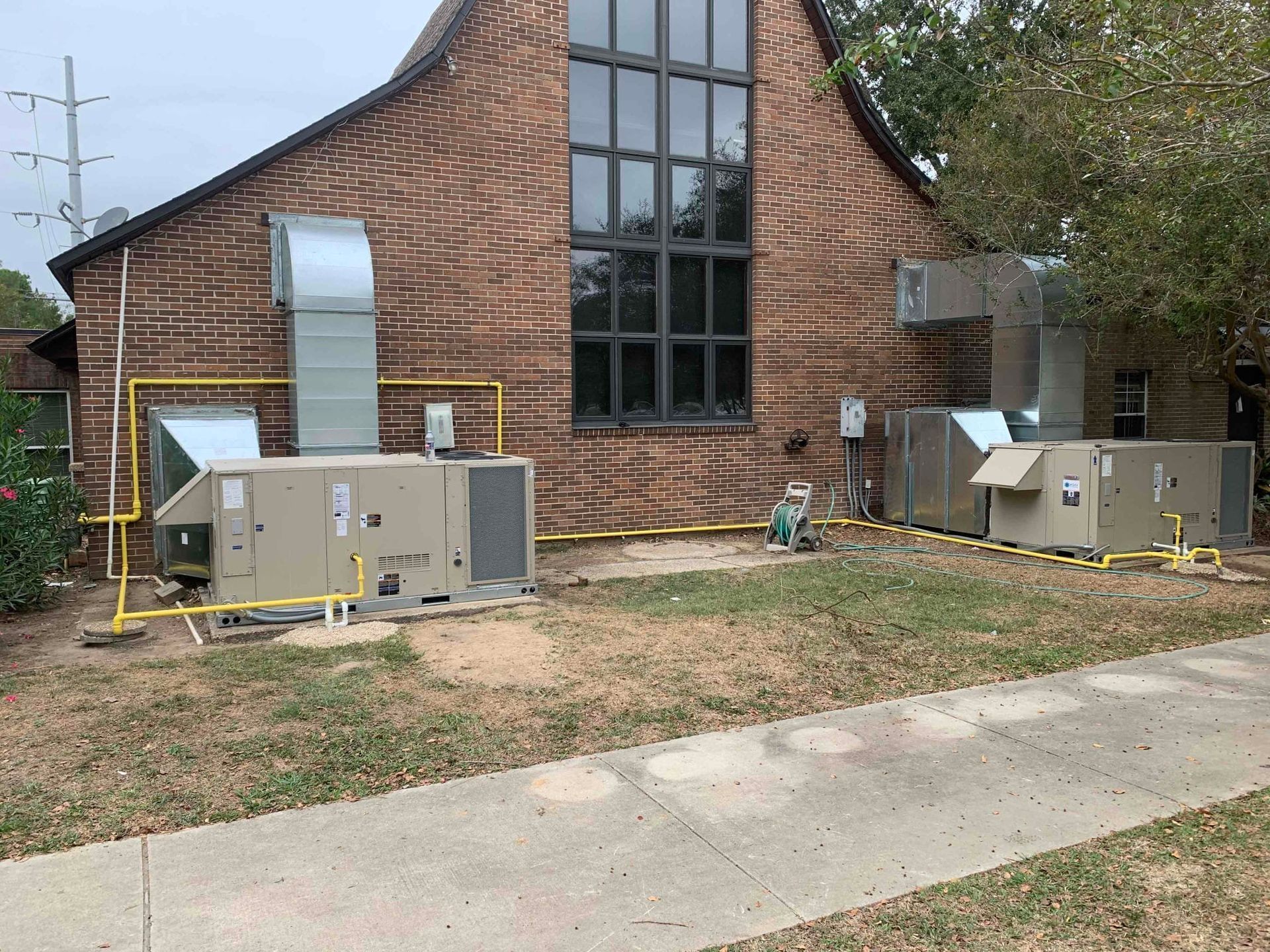 Brick building with glass tower and two outdoor HVAC units on a grassy lot beside a sidewalk