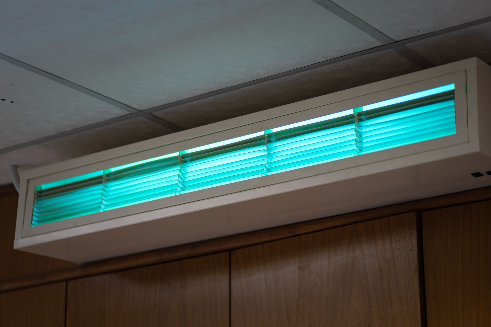Turquoise-lit wall-mounted heater or vent above wooden paneling and ceiling tiles