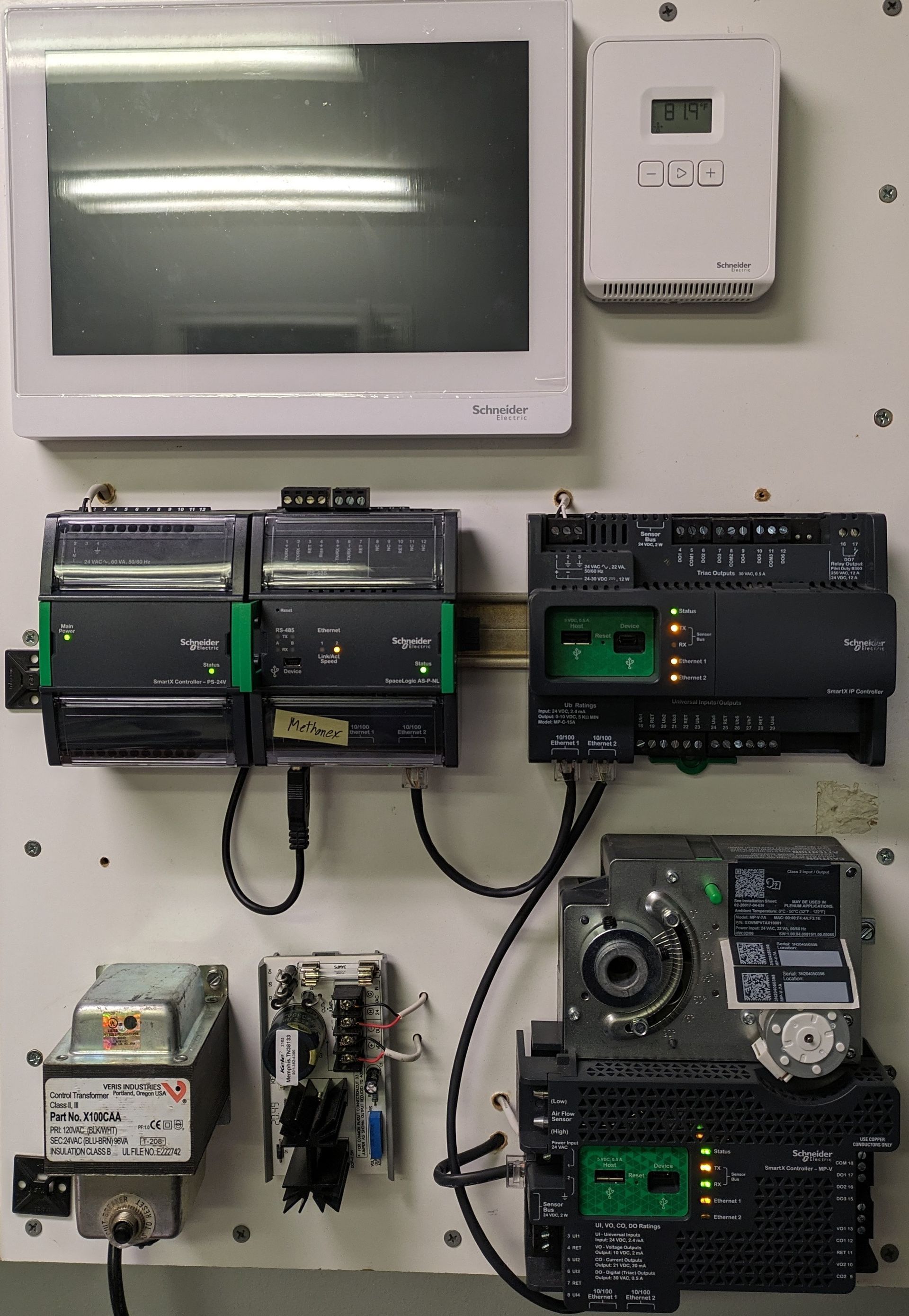 Wall-mounted industrial control panel with a monitor, thermostat, and several electrical modules with wiring.