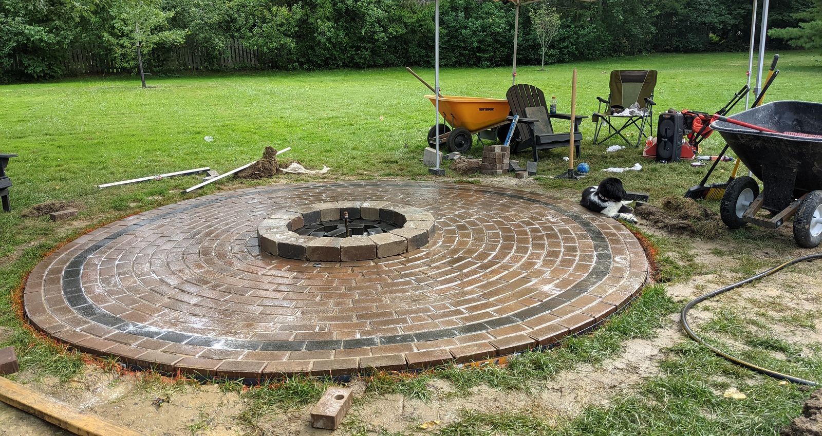Circular brick patio with fire pit, surrounded by tools and equipment in a grassy yard.