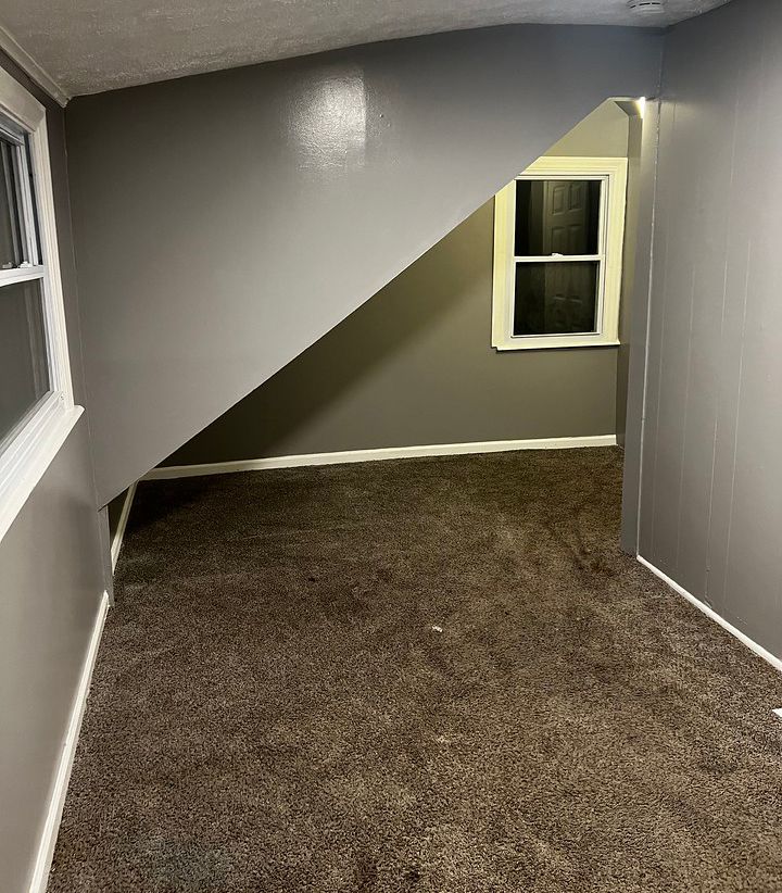 Room with gray walls, brown carpet, window, and space under a staircase.