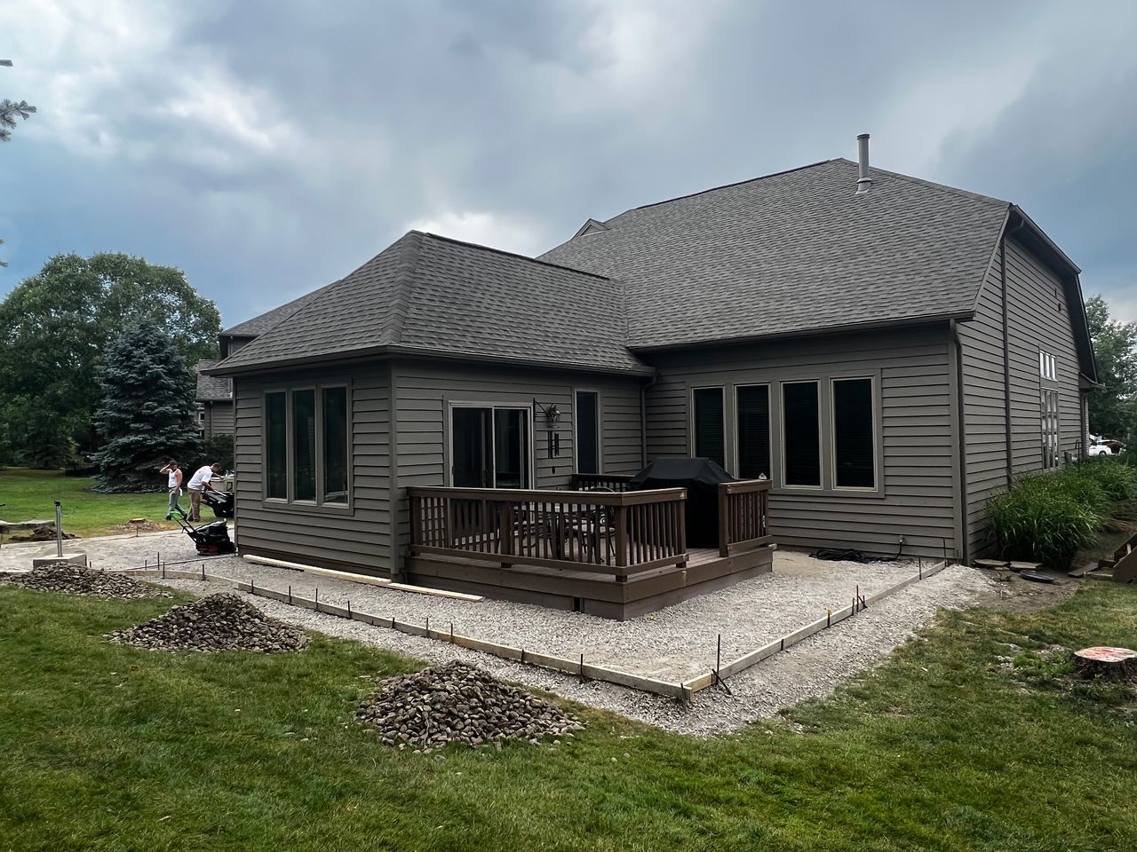 Backyard with home, deck, and patio under construction on a cloudy day. Gravel and concrete border are visible.