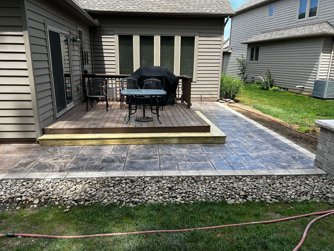 A back patio with a wooden deck, stamped concrete walkway, and rock border next to a lawn.