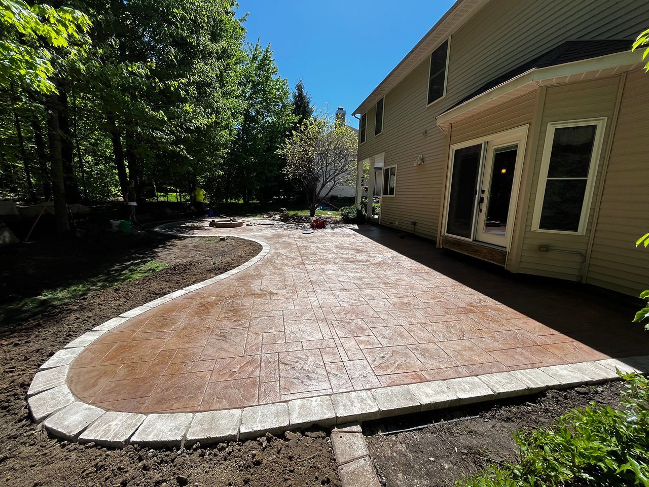 Stamped concrete patio with brick border, next to a two-story beige house and grassy area.