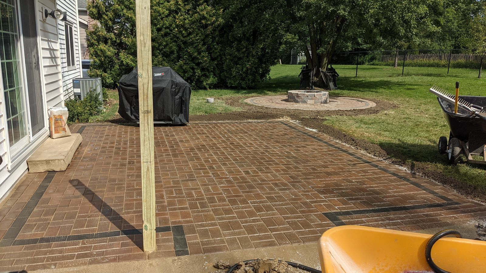 Brick patio with black border, barbecue grill, and wooden post, in a backyard setting.