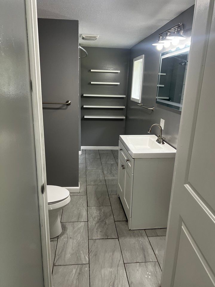 Bathroom with gray walls, white vanity and toilet, gray tiled floor, and open shelving.