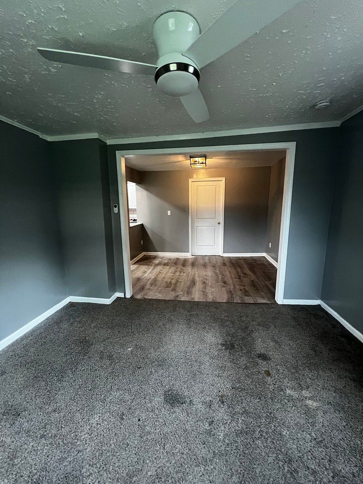 A room with gray walls and carpet, leading to another room with wood floors and a closed door.