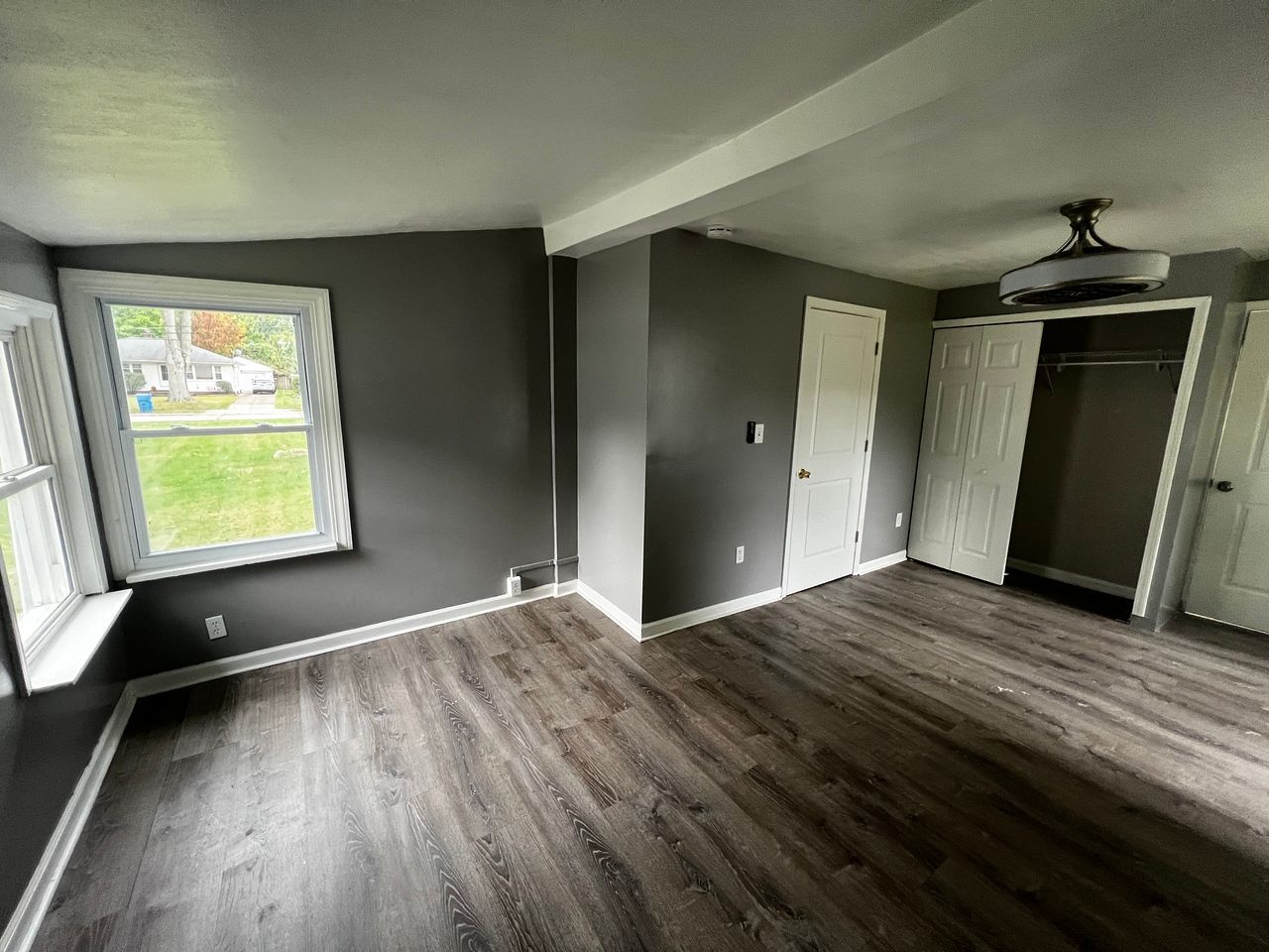 Gray-painted bedroom with wood-look floor, white trim, and a window overlooking a green lawn. Closet and a door are visible.