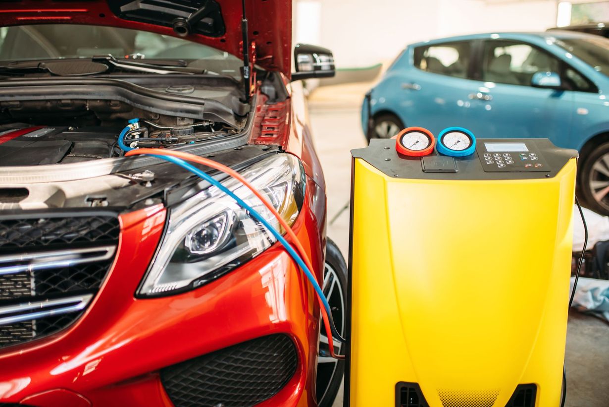 Red car with open hood beside yellow battery charger and another car in a garage.
