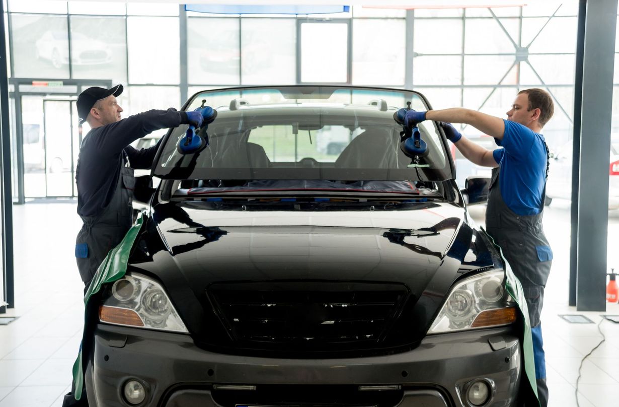 Two workers installing a windshield on a black car in a bright auto shop.