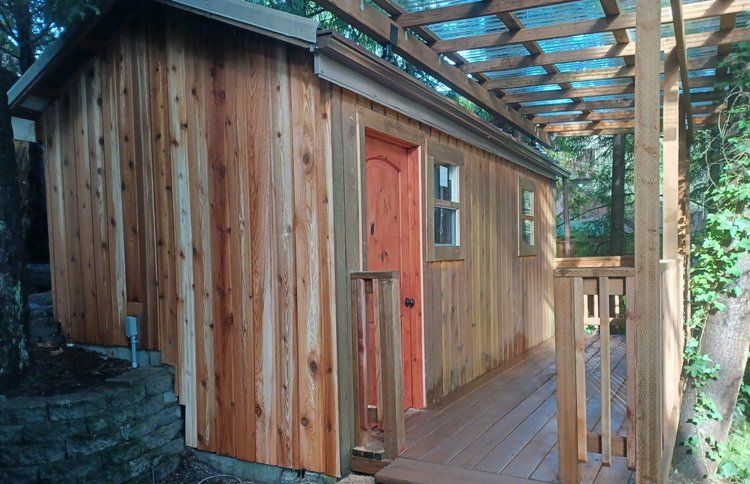 Wooden cabin with red door and porch, shaded by a pergola.