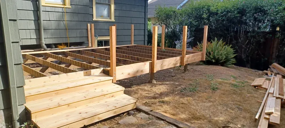 Wooden deck under construction next to a house with unfinished steps, dirt, and bushes.