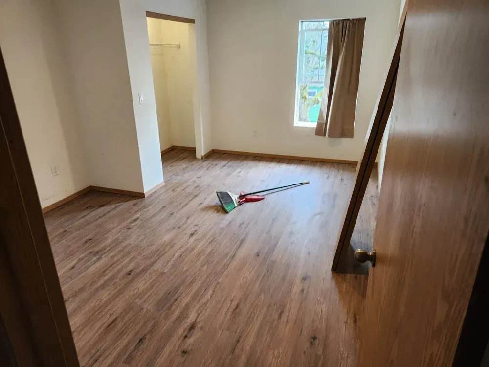 Empty bedroom with wood-look flooring, closet, window with curtain, and cleaning supplies.