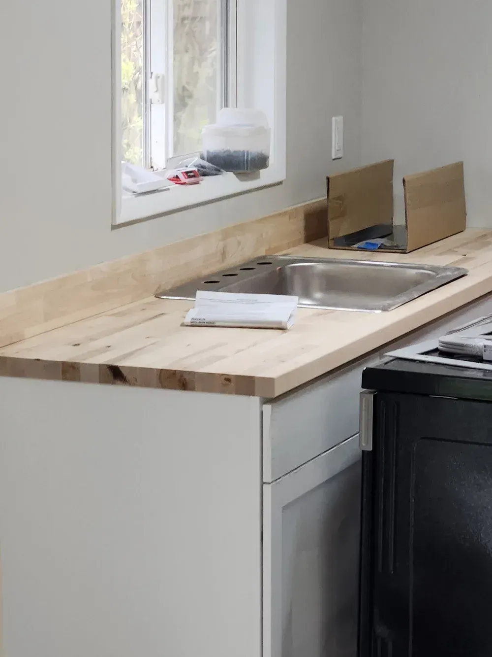 Kitchen countertop with sink, cardboard boxes, and unfinished wooden surface. White cabinets and a window are visible.