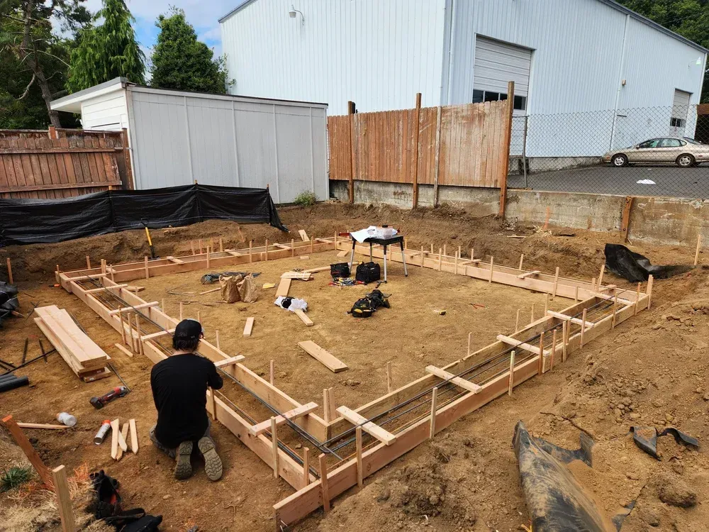 Person kneeling, building wooden frame for a concrete foundation in a dirt yard.