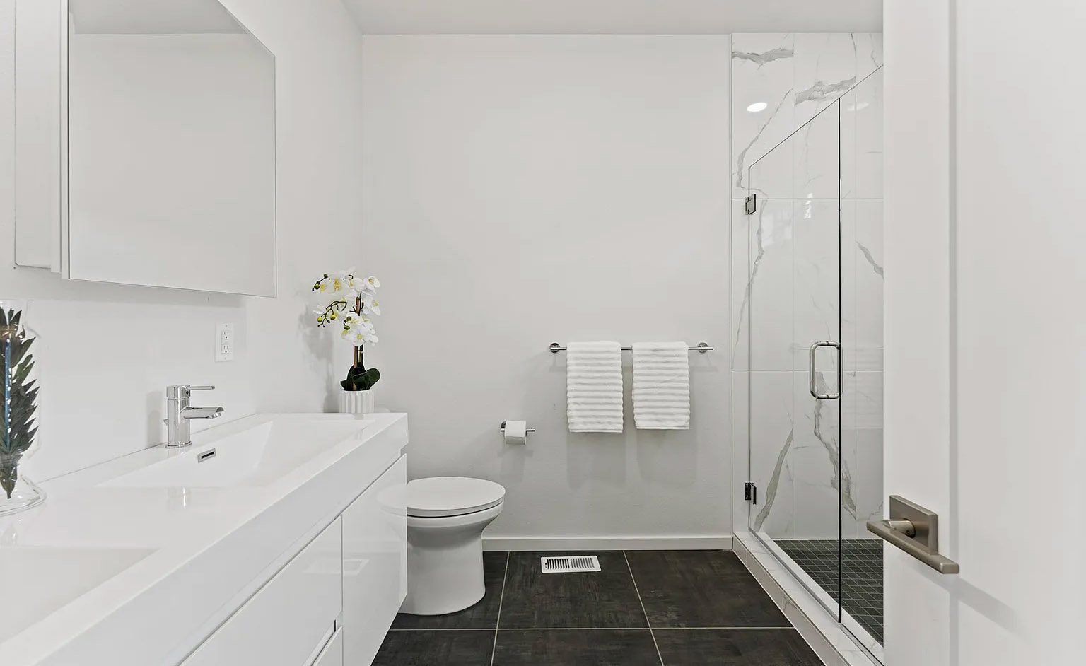 White bathroom with floating vanity, toilet, glass shower, and dark tile floor.