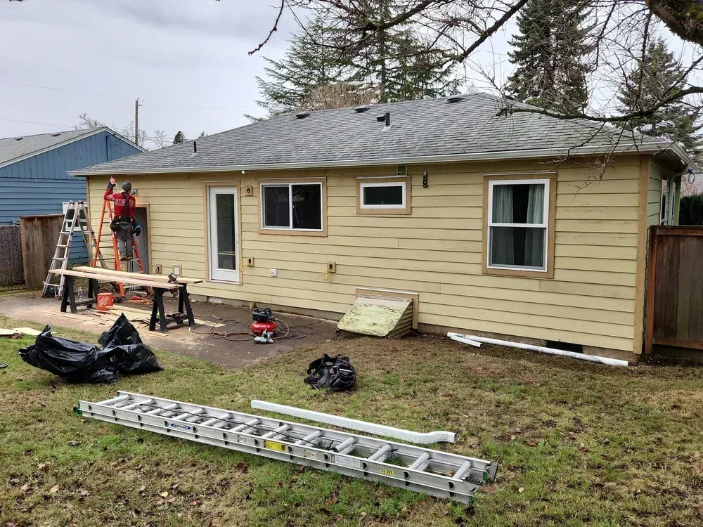 A person on a ladder repairs the siding of a yellow house with a gray roof; construction tools are on the ground.